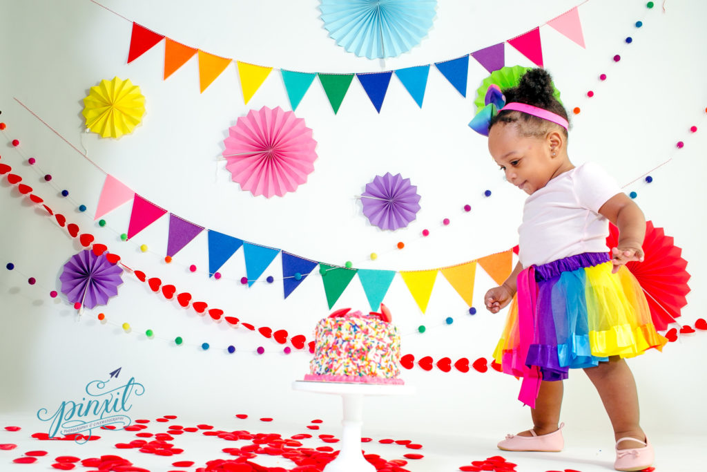 little girl wearing a rainbow tutu staring at a sprinkle covered smash cake  in front of a white back drop covered in rainbow decor 
