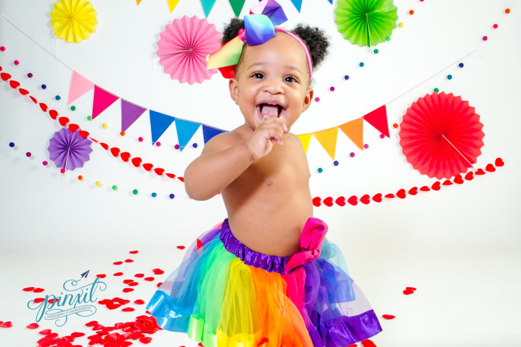 little girl wearing a rainbow tutu running around in front of a white back drop covered in rainbow decor 