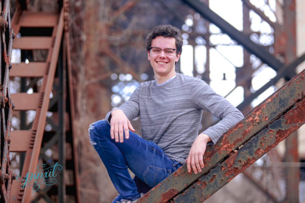 teen boy wearing glasses, grey long sleeve t shirt and blue jeans sitting on rusted urban bridge legs 