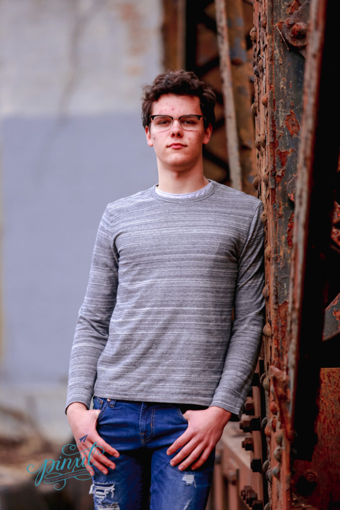 teen boy wearing glasses, grey long sleeve t shirt and blue jeans leaning on rusted urban bridge legs 