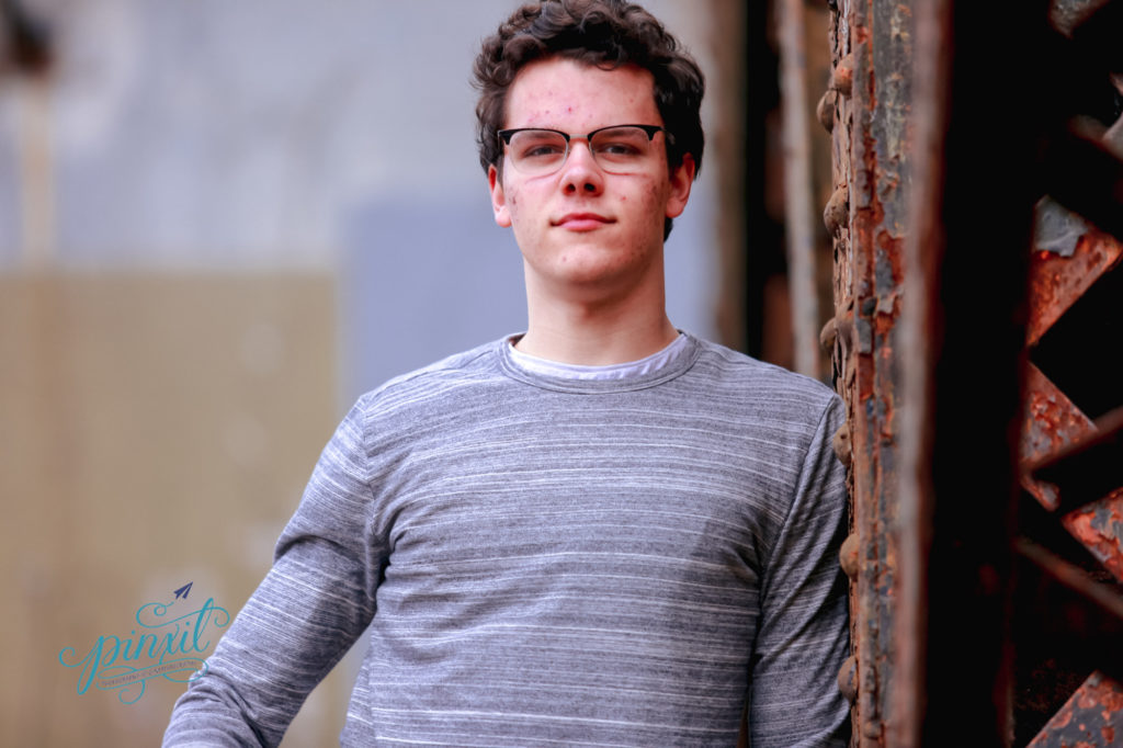 teen boy wearing glasses, grey long sleeve t shirt and blue jeans leaningon rusted urban bridge legs 