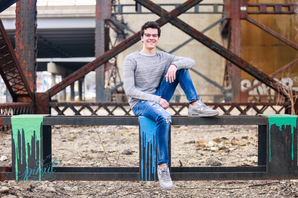 teen boy wearing glasses, grey long sleeve t shirt and blue jeans sitting on rusted urban bridge legs with green paint on each edge 