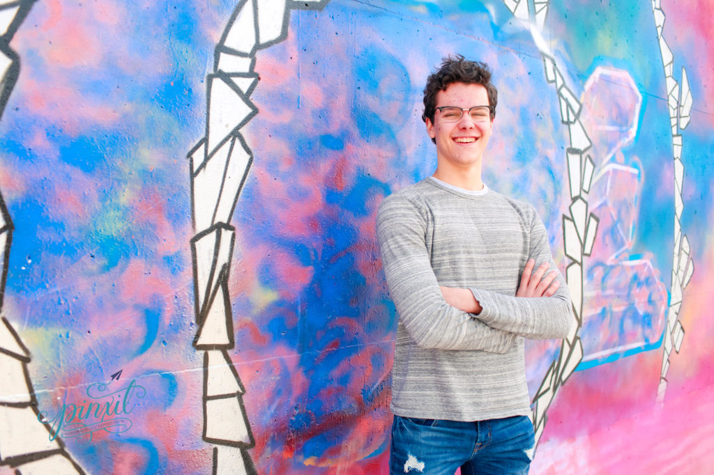 teen boy wearing glasses, grey long sleeve t shirt and blue jeans leaning on a pink and blue graffiti painted  wall with white accents  
