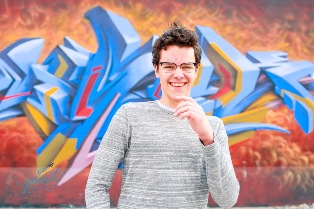 teen boy wearing glasses, grey long sleeve t shirt and blue jeans laughing in front of a orange yellow and blue graffiti painted  wall with white accents  