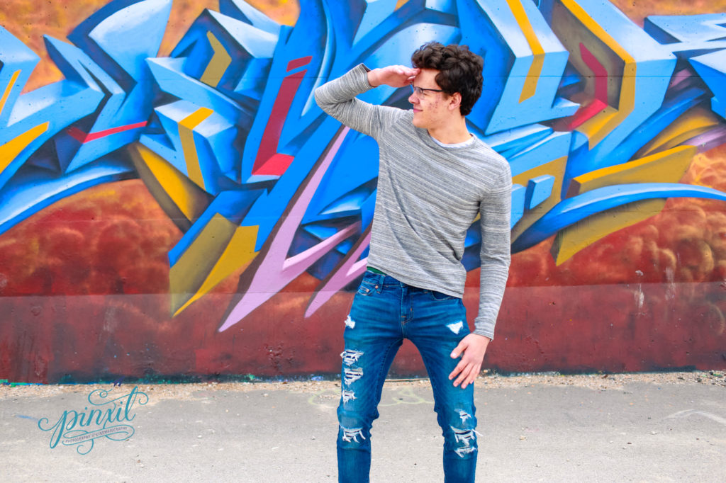 teen boy wearing glasses, grey long sleeve t shirt and blue jeans laughing in front of a orange yellow and blue graffiti painted  wall with white accents 