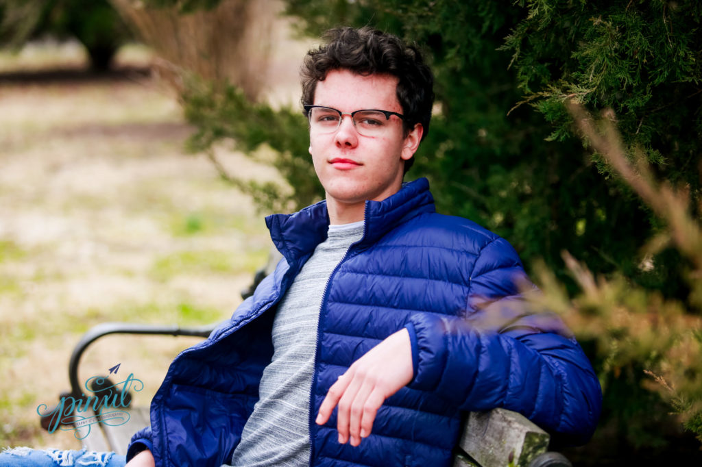 St Louis high school senior wearing glasses and blue puffy coat leaning back on a park bench in tower grove park 