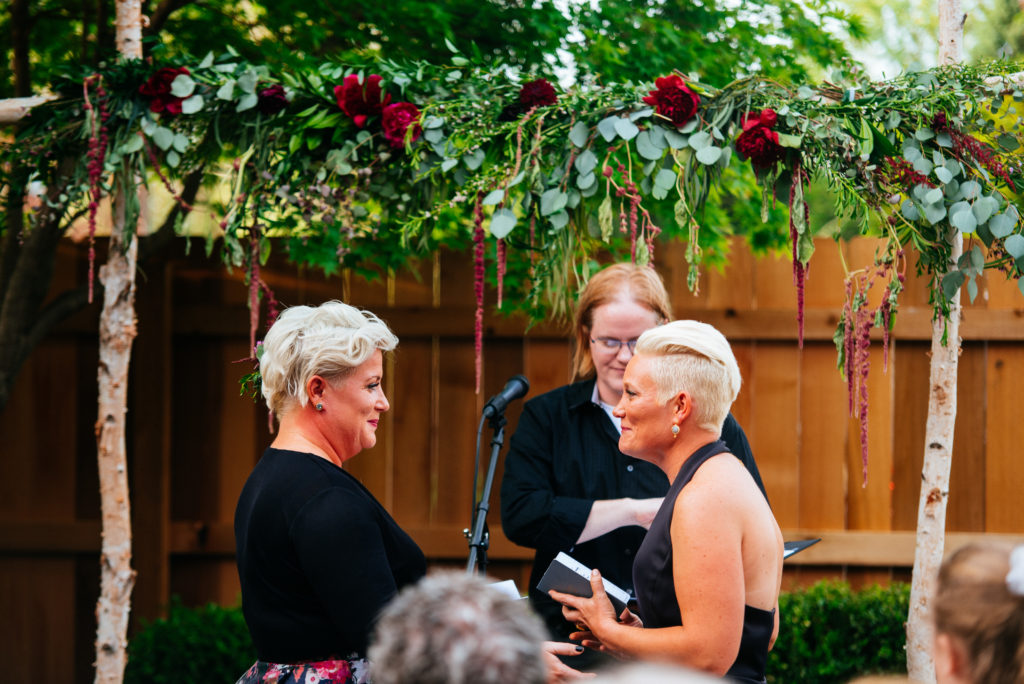 female couple stands under a beautiful red rose trellis gazing at each other as their ceremony starts. Their Chosen officiant is preparing to speak behind them. 