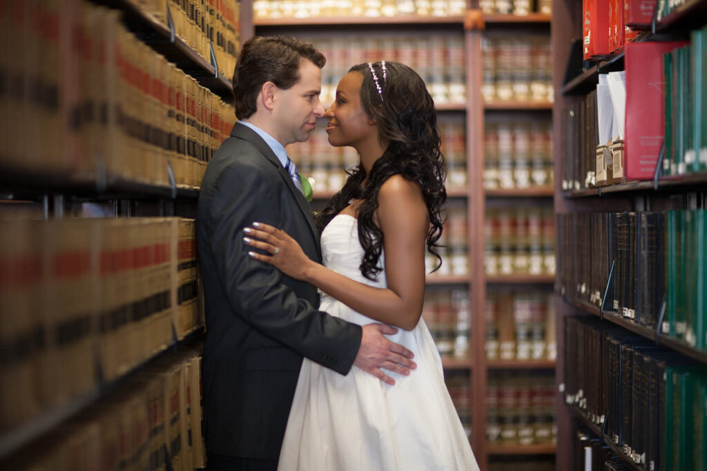 An interracial Bride and groom stand in the book stacks of the SLU law library in St.Louis, gazing into each others eyes.