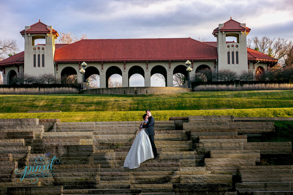 Groom hugs the bride tightly standing on stones steps with the worlds fair pavilion in Forest Park, St. Louis in the background