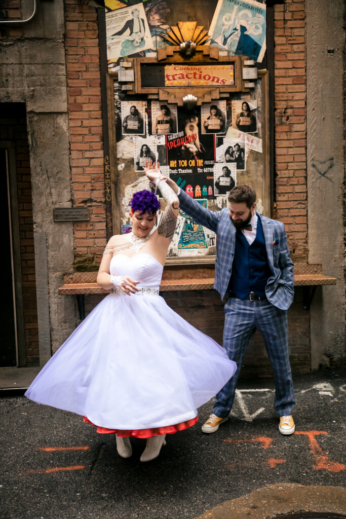 a couple dancing behind the Thaxton Speakeasy where they had their wedding in St. Louis, She's got rainbow hair and rainbow colors under her white wedding dress and vintage white shoes and hes wearing a blue plaid suit with converse against a colorful mural wall, alternative St. Louis wedding portrait